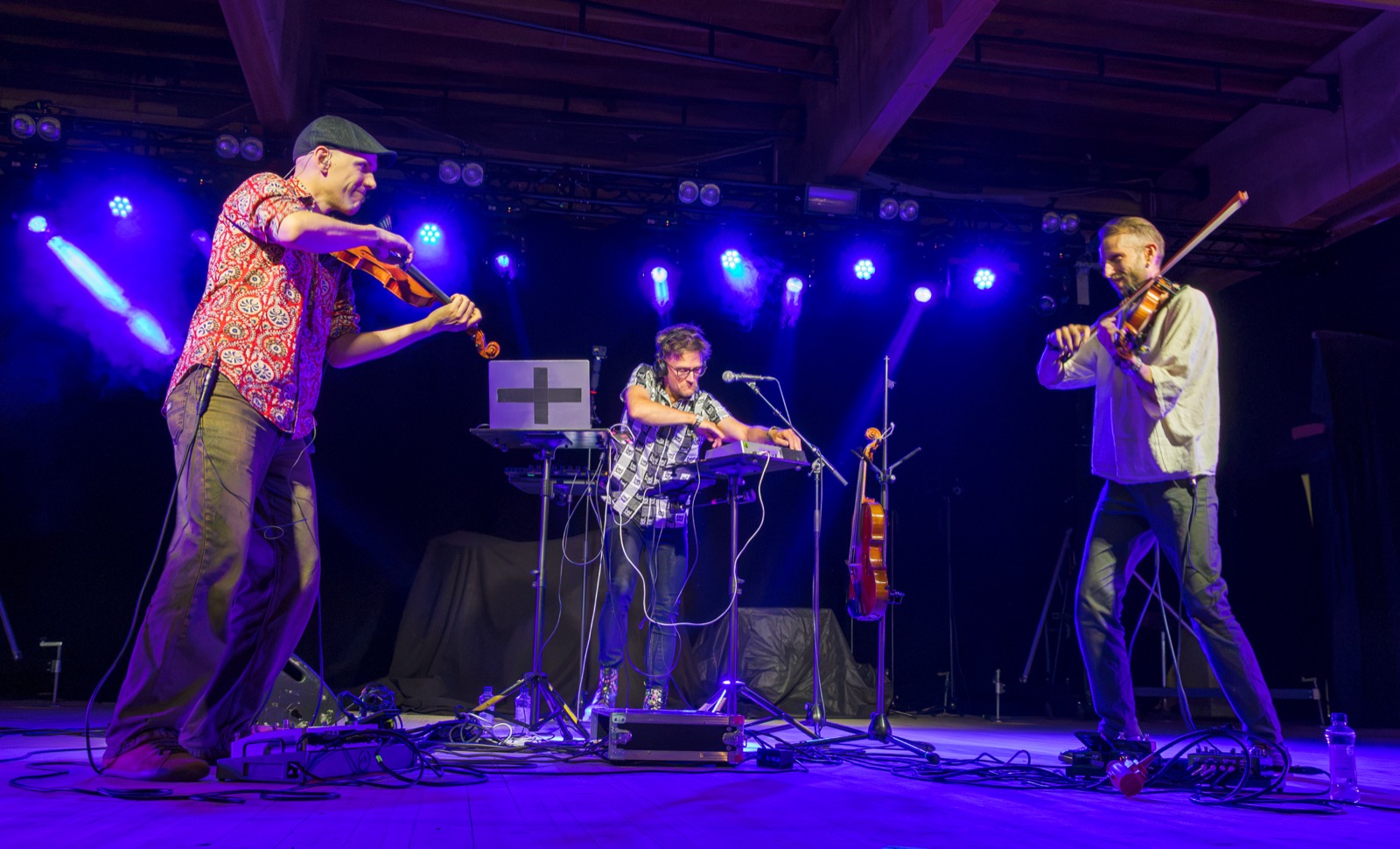 Tero Hyväluoma and Esko Järvelä playing fiddles on a blue-lit stage, Antti Puumalainen at the electronics between them